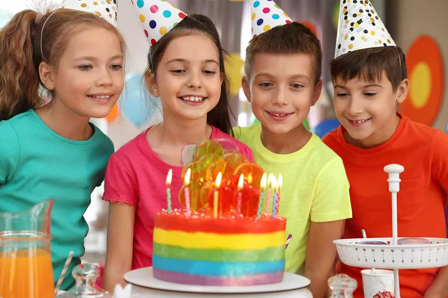 Kids in party hats around a rainbow birthday cake
